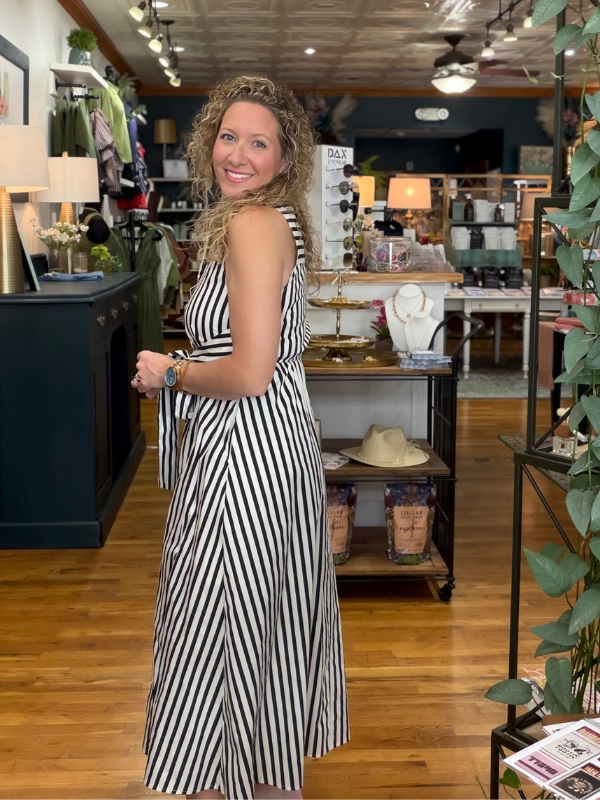 A woman with curly blonde hair stands in a boutique shop wearing a sleeveless black and white vertically striped midi dress. She is smiling and looking back over her shoulder, with store fixtures, wood floors, and plants visible in the background.