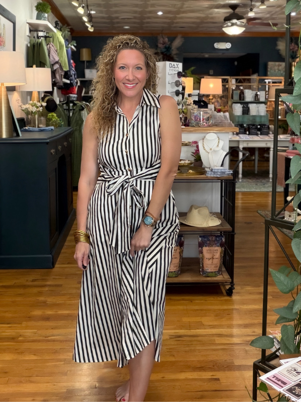 A woman with curly blonde hair stands in a boutique shop wearing a sleeveless black and white vertically striped midi dress with a tie waist. The background shows wooden floors, store fixtures, and other merchandise.