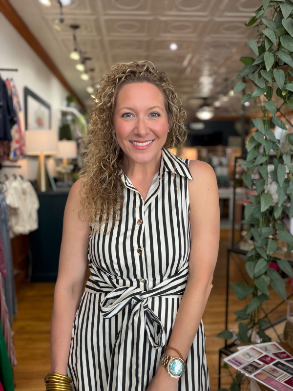 A smiling woman with curly blonde hair wearing a sleeveless black and white vertically striped belted dress, standing inside a boutique store.