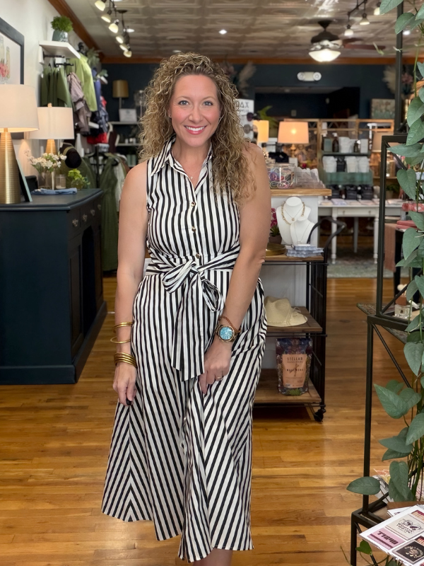 A woman with curly blonde hair stands in a boutique shop wearing a sleeveless black and white vertically striped midi dress with a tie waist. The background shows wooden floors, clothing racks, and store fixtures.