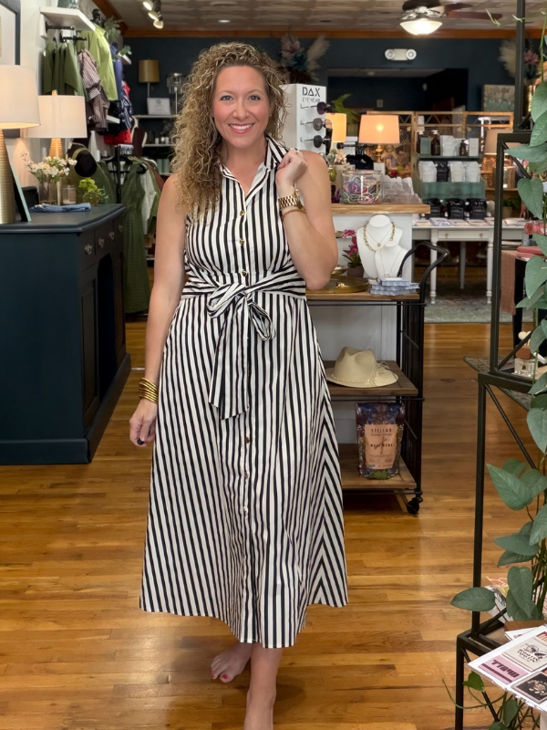 A woman with curly blonde hair stands barefoot in a boutique shop wearing a sleeveless black and white vertically striped midi dress with a tie waist. The background shows wooden floors, clothing racks, and store fixtures.