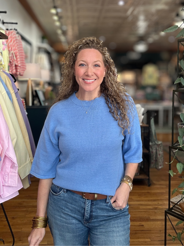 Woman wearing a blue sweater and jeans standing in a store with clothing racks in the background.