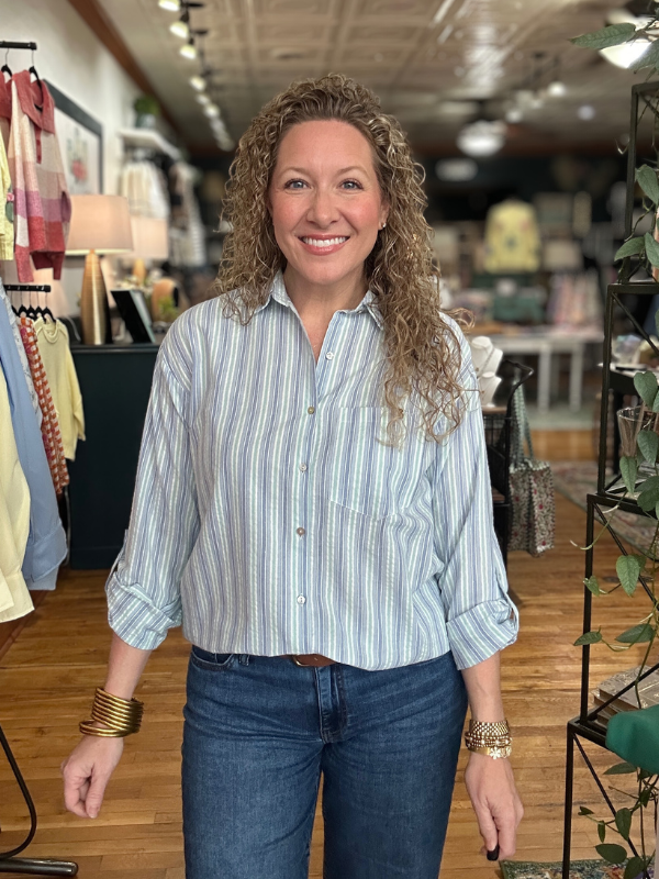 Woman in a striped shirt and jeans standing in a store with clothing racks and decor in the background.
