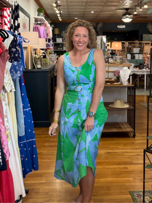 Woman in a green and blue dress standing in a store with racks of clothes in the background.