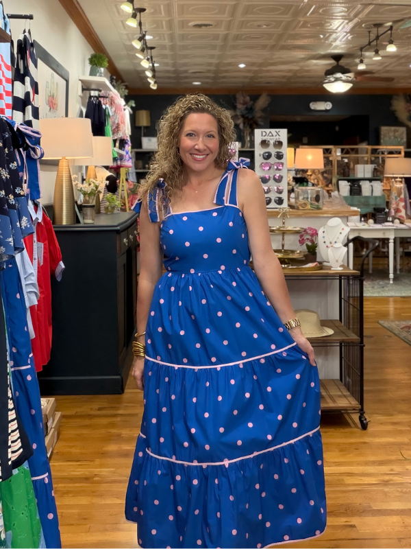 Woman in a blue polka dot dress standing in a store with clothing racks and displays.