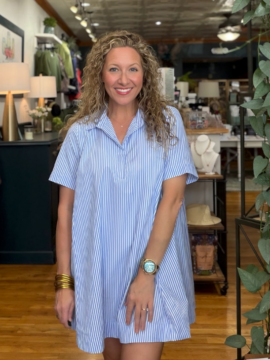 A smiling woman with curly hair stands in a boutique wearing a light blue and white vertically striped short-sleeve shirt dress. She is accessorized with gold bracelets, a watch, and rings, with wooden floors and store fixtures visible in the background.