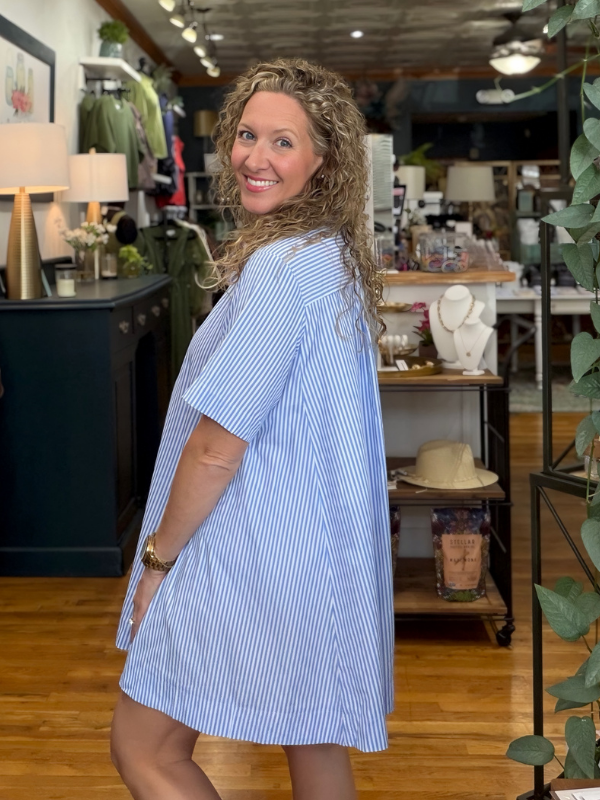 A woman with curly blonde hair wearing a light blue and white vertically striped short-sleeve shift dress, posing inside a boutique shop.
