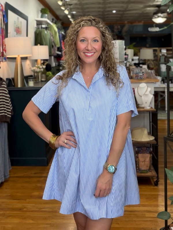 A smiling woman with curly blonde hair stands in a boutique wearing a light blue and white vertically striped short-sleeve shirt dress. The dress has a pointed collar and a flared skirt.
