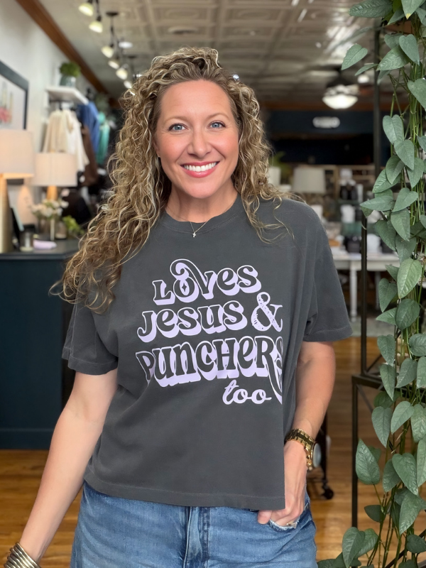 A smiling woman with curly blonde hair wearing a dark gray graphic t-shirt that reads 'Loves Jesus & Punchers too' in white and purple lettering, paired with blue jeans, standing inside a retail store.