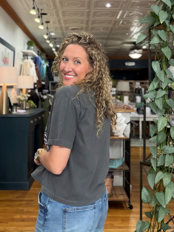 A woman with curly blonde hair wearing a dark gray t-shirt and blue jeans, looking back over her shoulder in a retail store setting.