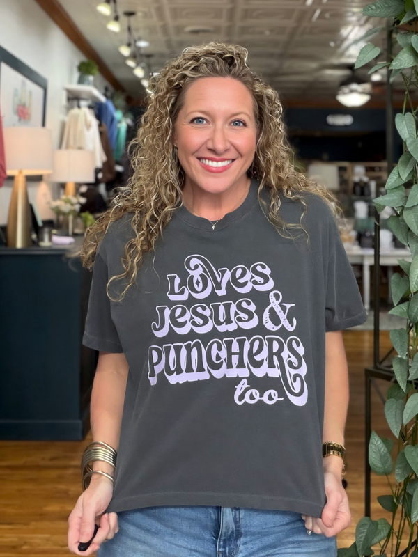 A smiling woman with curly blonde hair stands in a retail store wearing a dark charcoal gray graphic t-shirt with the text 'Loves Jesus & Punchers too' in a light lavender-purple stylized font, paired with blue jeans.
