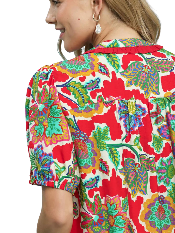 A close-up side view of a woman wearing a short-sleeve blouse with a vibrant, multicolored floral and paisley print on a cream background. The blouse features bold red, green, blue, and yellow patterns.