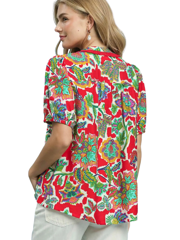 A woman shown from the back wearing a short-sleeve button-up shirt with a vibrant red, green, and blue tropical floral pattern, paired with light-colored pants, against a plain white background.