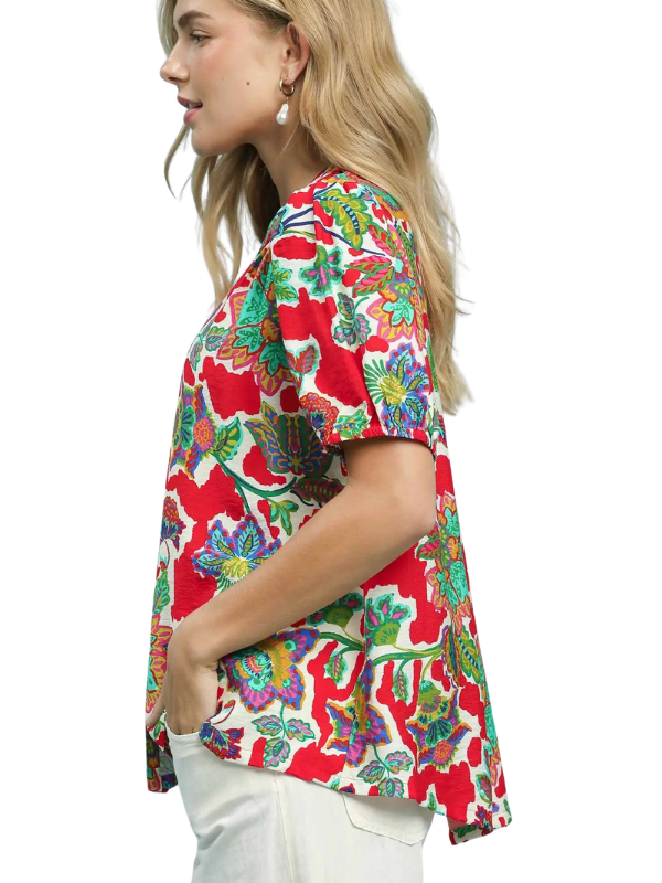 A woman shown in side profile wearing a short-sleeve button-up blouse with a bold red, green, and blue floral pattern, paired with white pants, against a plain white background.