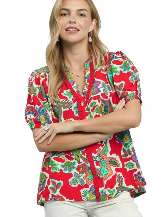 A woman wearing a vibrant red short-sleeve blouse with a colorful tropical floral and bird print, paired with light beige pants, set against a plain white background.