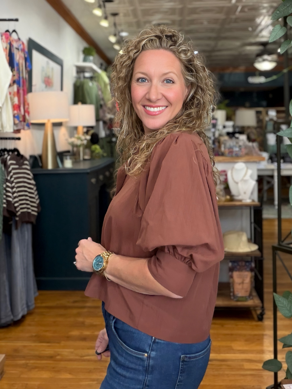 A smiling woman with curly blonde hair stands in a boutique shop, wearing a rust-brown puff-sleeve blouse and blue jeans.