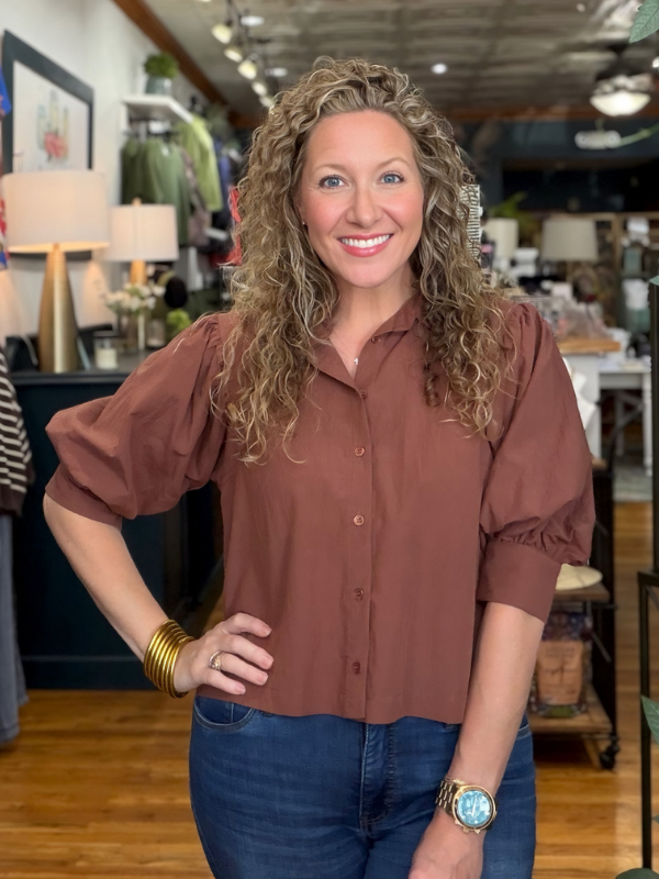 A smiling woman with curly blonde hair wearing a rust-brown puff-sleeve button-up blouse and blue jeans, standing in a retail store.