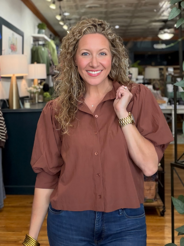A smiling woman with curly blonde hair wearing a rust-brown short-sleeve button-up blouse and blue jeans, standing in a retail store interior.