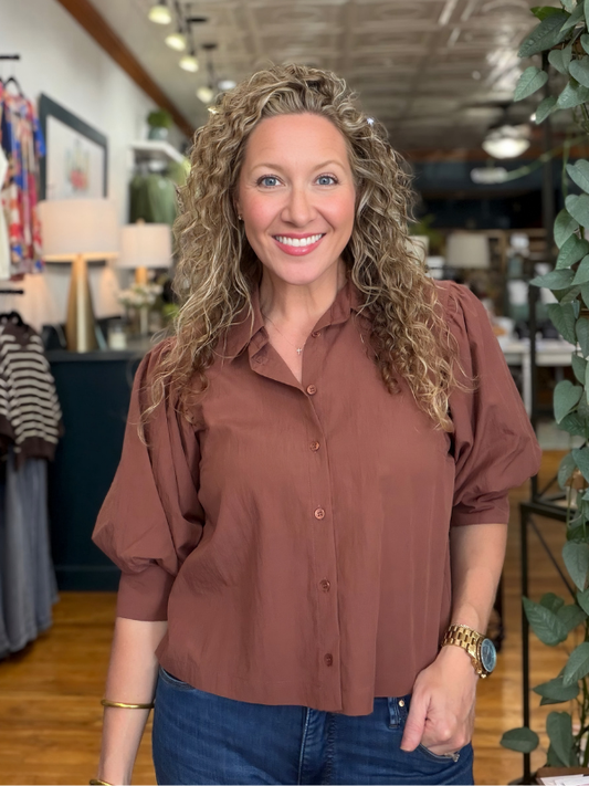 A smiling woman with curly blonde hair wearing a rust-brown button-down blouse and blue jeans, standing inside a retail store.