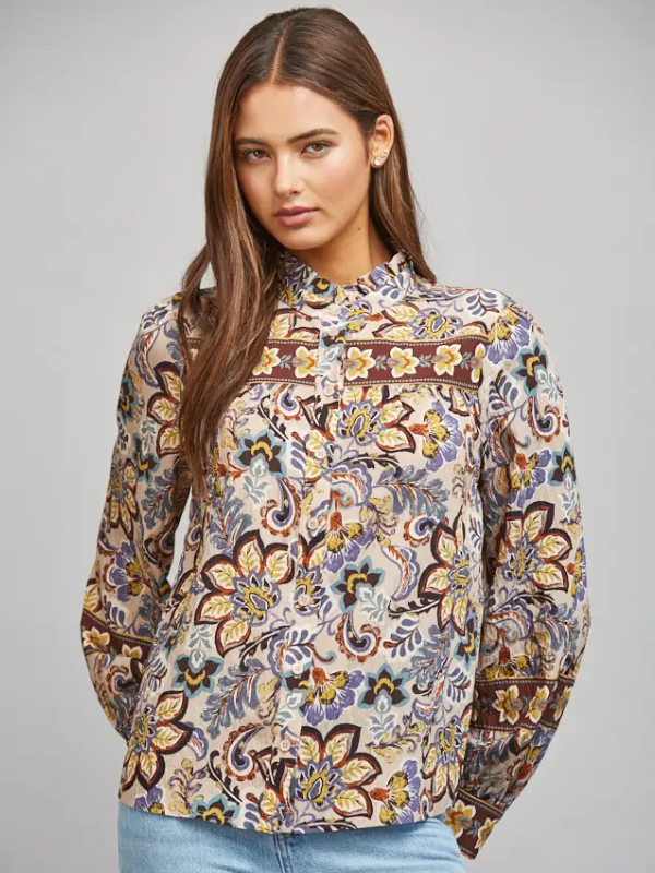A woman modeling a long-sleeve button-up blouse with a detailed multicolor floral and paisley print in brown, yellow, blue, and beige tones, paired with light blue jeans against a solid gray studio background.