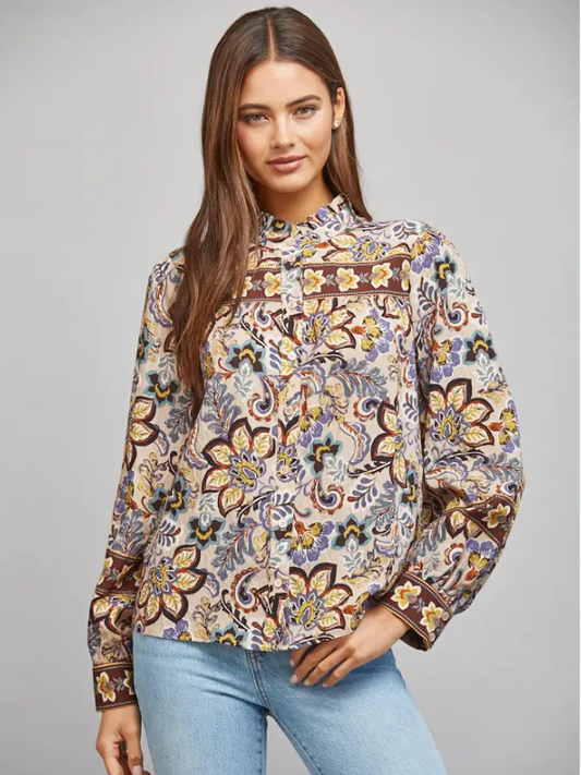 A woman modeling a long-sleeve high-neck blouse with a colorful floral and paisley print, paired with light blue jeans, against a solid gray studio background.