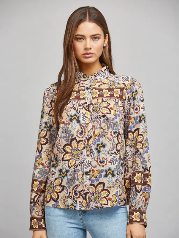 A woman modeling a long-sleeve high-neck blouse with an intricate multicolor floral and paisley print in beige, brown, yellow, and blue, paired with light blue jeans against a solid gray studio background.