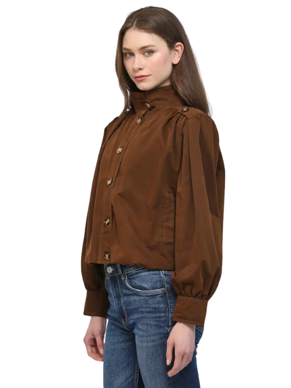 A woman modeling a brown long-sleeve button-up jacket with gathered shoulders and a high collar, paired with blue jeans, against a plain white background.