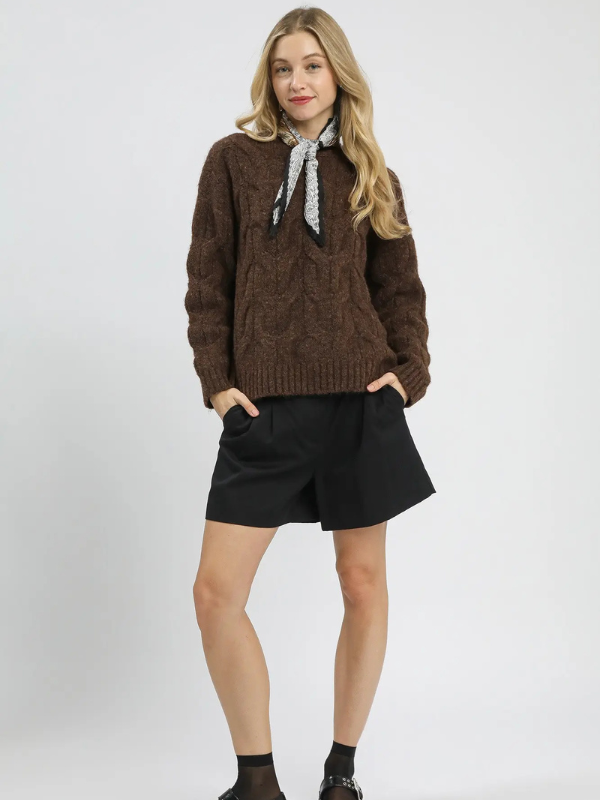 Woman wearing a brown cable knit sweater with a black collar and black shorts on a white background