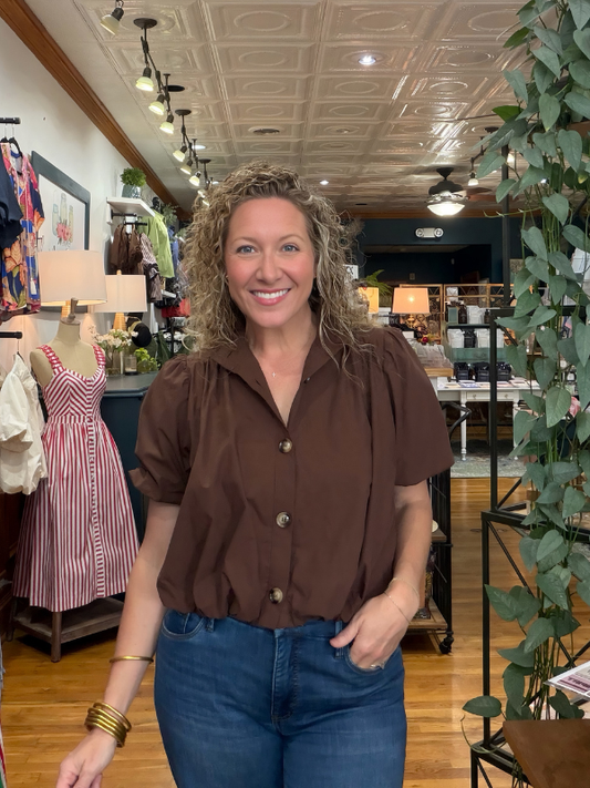 A smiling woman with curly blonde hair stands in a boutique shop wearing a brown short-sleeve button-up blouse and blue jeans.