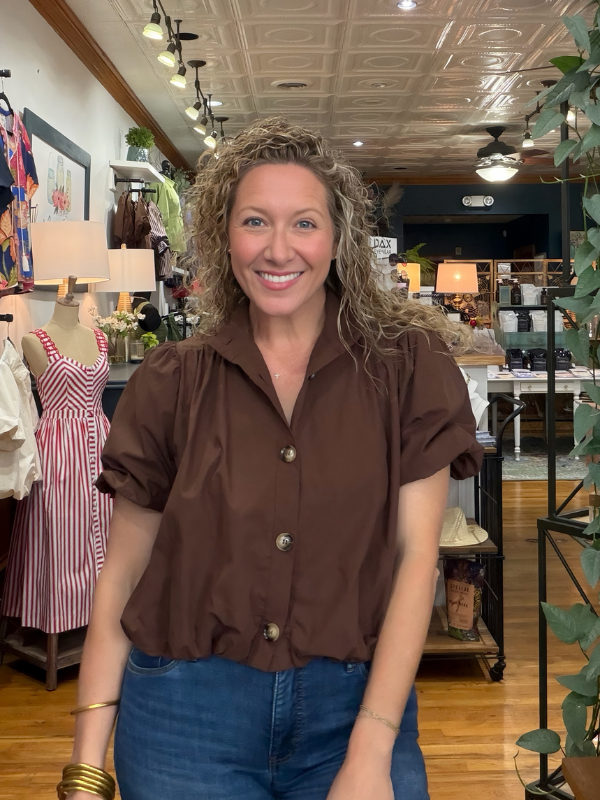A smiling woman with curly hair stands in a boutique shop wearing a brown short-sleeve button-up shirt and blue jeans.
