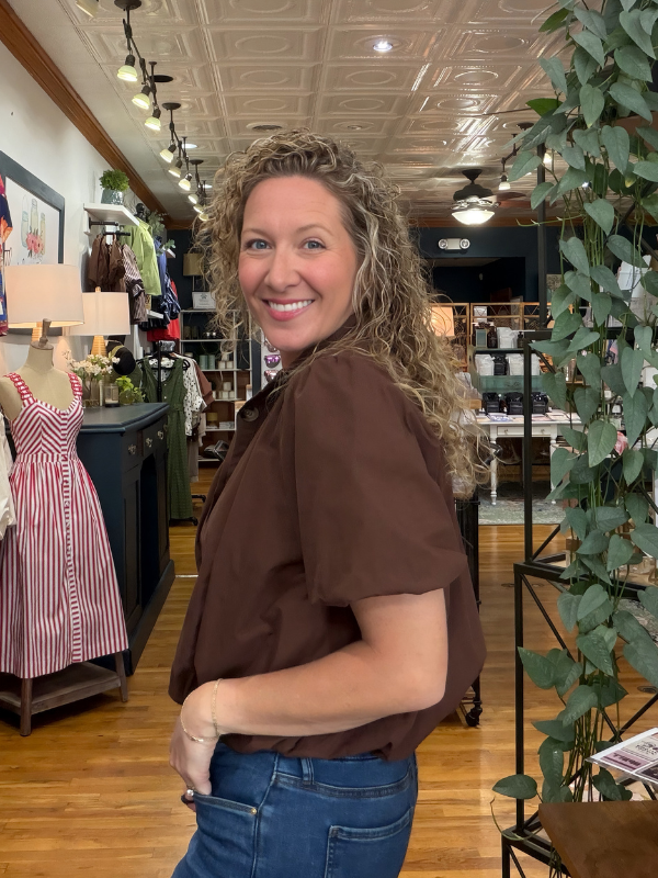A woman with curly blonde hair smiling and looking back over her shoulder inside a boutique. She is wearing a brown short-sleeve button-up shirt and blue jeans.
