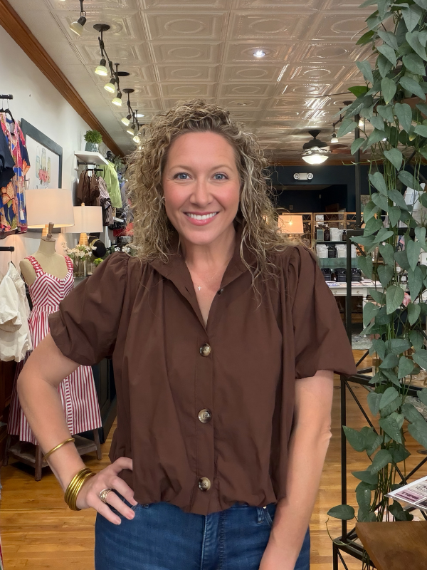 A smiling woman with curly blonde hair stands in a boutique wearing a brown short-sleeve button-up shirt and blue jeans.