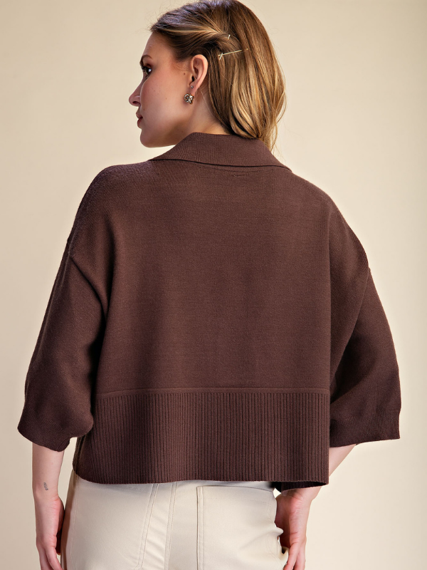 A woman shown from the back wearing a cropped brown knit sweater with three-quarter sleeves and a ribbed hem, paired with off-white pants, against a plain beige studio background.