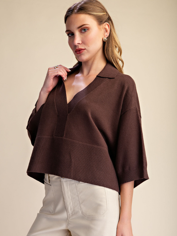 A woman modeling a brown knit polo-style top with wide three-quarter sleeves and a ribbed hem, paired with light beige pants, against a plain beige studio background.