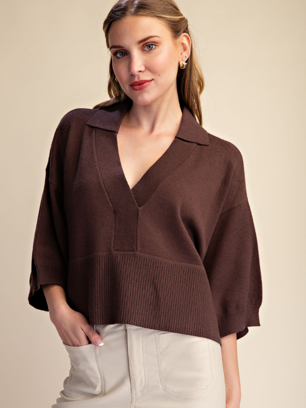 A woman wearing a dark brown knit polo sweater with a V-neck and wide three-quarter sleeves, paired with off-white pants, against a solid beige background.