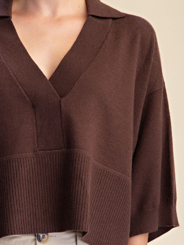 A close-up of a person wearing a dark brown knit top with a V-neck, wide three-quarter sleeves, and a ribbed hem, paired with light-colored pants against a plain beige background.