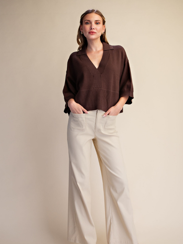 A woman modeling a dark brown V-neck blouse with three-quarter sleeves and cream wide-leg trousers, standing against a solid beige studio background.