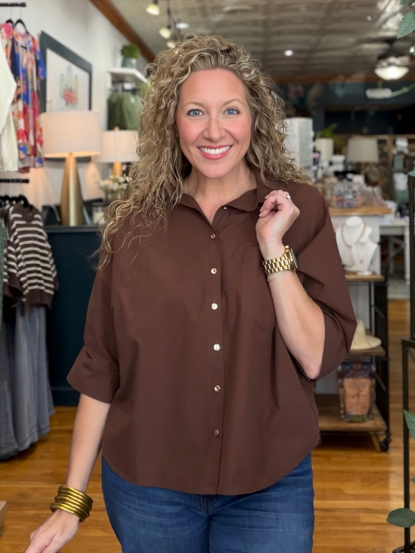 A smiling woman with curly blonde hair wearing a brown short-sleeve button-up shirt and blue jeans, standing inside a retail store.