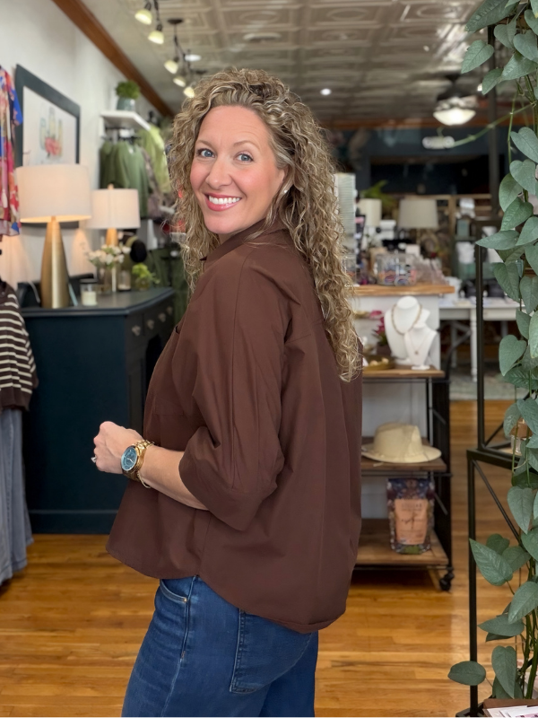 A smiling woman with curly blonde hair wearing a brown long-sleeve blouse and blue jeans, standing in a retail store interior.