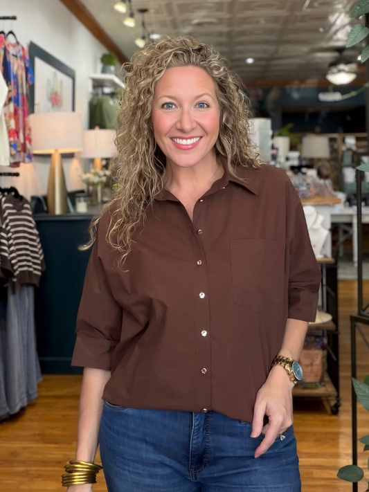 A smiling woman with curly blonde hair wearing a brown button-down shirt and blue jeans, standing inside a retail store.