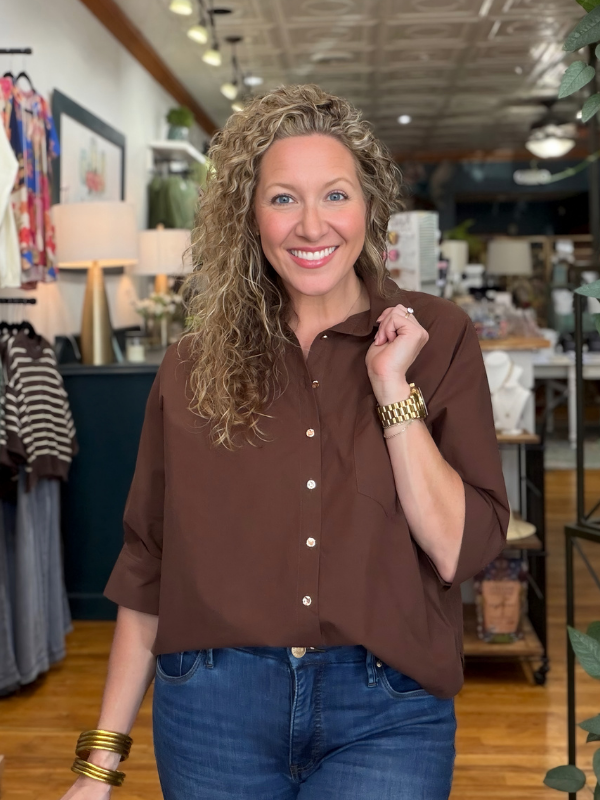 A smiling woman with curly blonde hair stands in a boutique wearing a brown button-up shirt and blue jeans, with gold bracelets on her wrists.