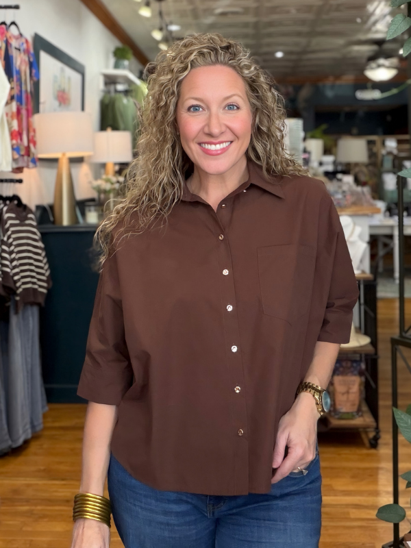 A smiling woman with curly blonde hair wearing a brown button-down shirt and blue jeans, standing inside a retail store.