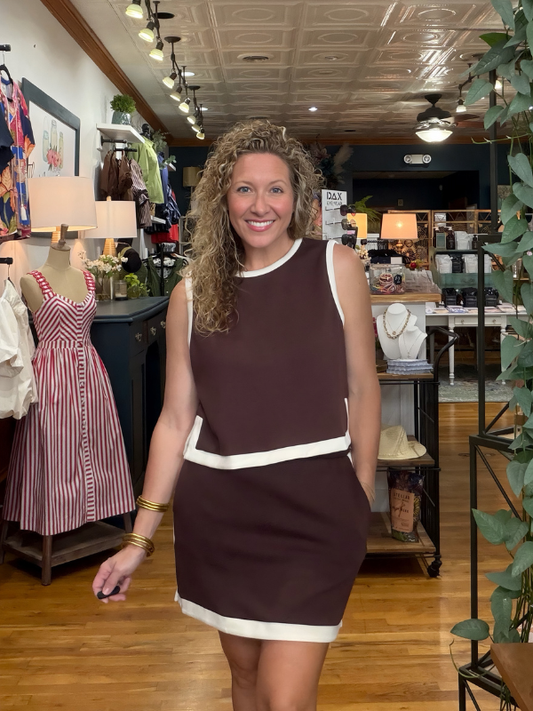 A woman with curly hair stands in a boutique wearing a sleeveless, knee-length dress in dark brown with white contrast trim. The dress features a crew neck, a horizontal white band at the waist, and a white hem.
