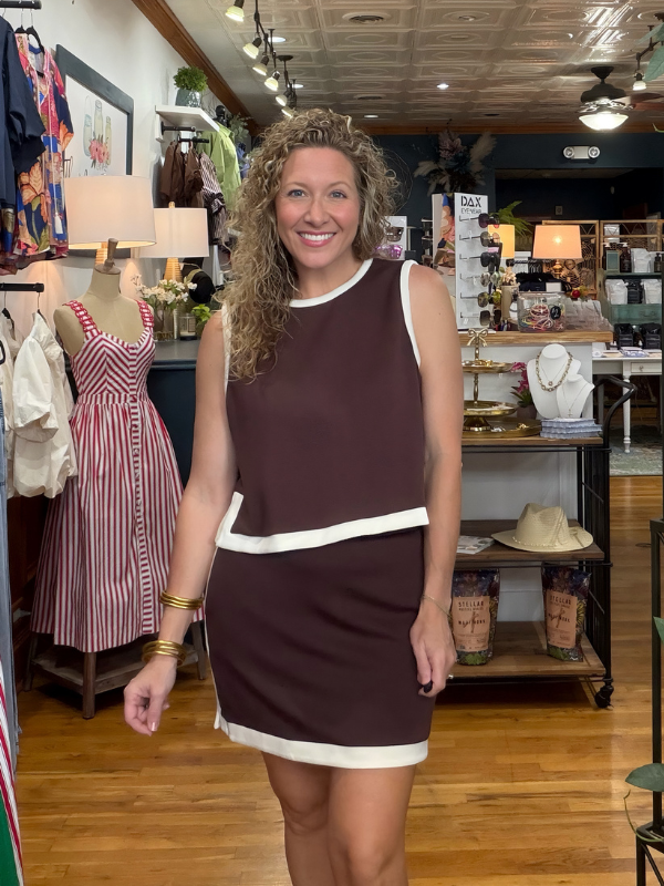 A woman with curly hair stands in a boutique wearing a sleeveless brown mini dress with white contrast trim. The dress features a crew neck, dropped shoulders, a horizontal white band at the waist, and a white hem.