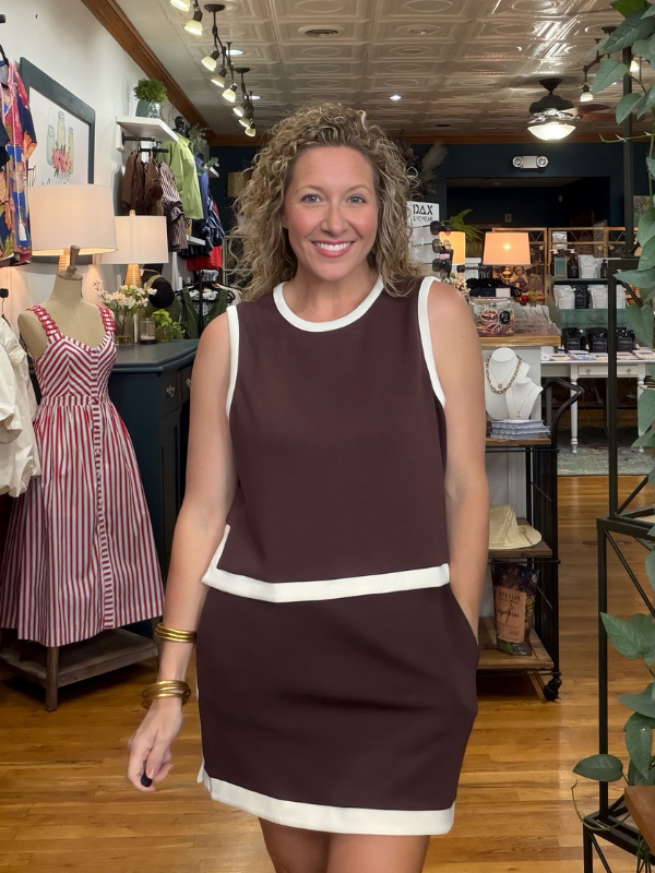 A smiling woman with curly hair stands in a clothing boutique wearing a sleeveless brown dress with white contrast trim at the neckline, armholes, hem, and a horizontal seam. Mannequins and store fixtures are visible in the background.