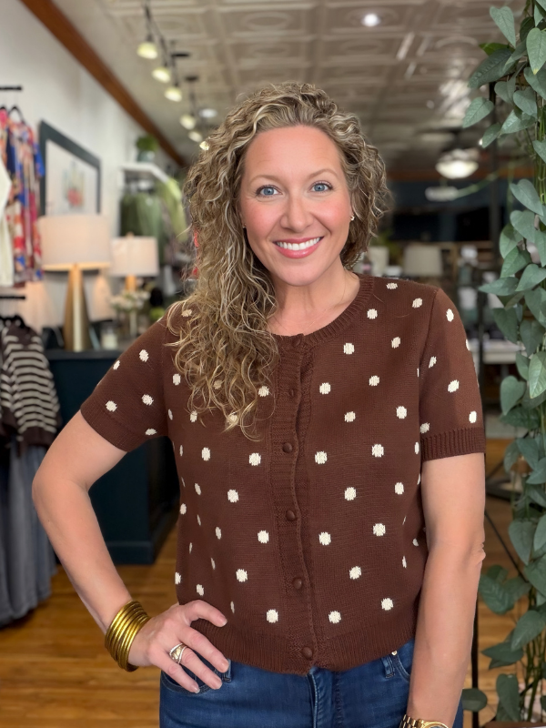 A smiling woman with curly blonde hair wearing a brown short-sleeve knit cardigan with white polka dots, standing in a retail store.