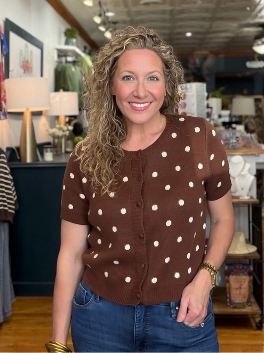 A smiling woman with curly blonde hair wearing a brown short-sleeve cardigan with white polka dots and blue jeans, standing inside a retail store.