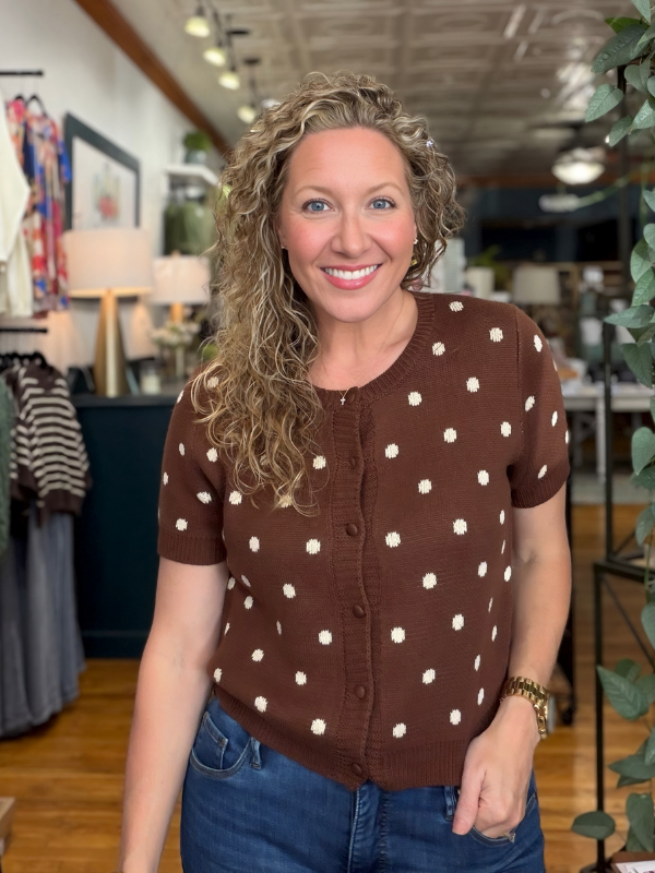 A smiling woman with curly blonde hair wearing a brown short-sleeve knit top with white polka dots and blue jeans, standing inside a boutique store.