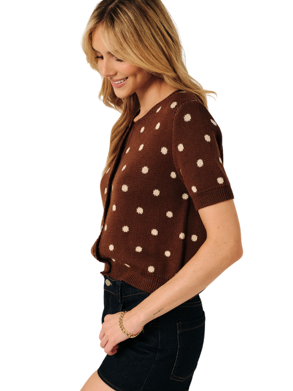 A woman wearing a brown short-sleeve knit top with cream polka dots, paired with dark denim shorts, shown from a side profile against a plain white background.