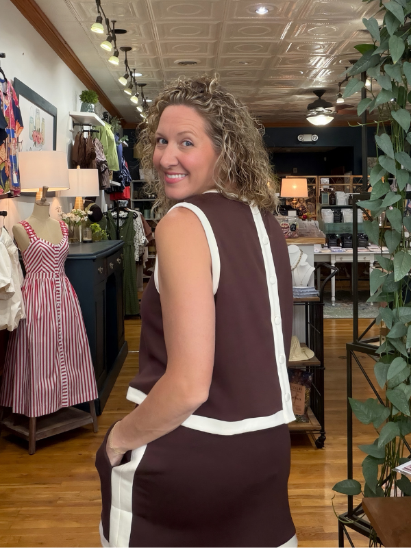 A woman wearing a sleeveless brown dress with white contrast trim, photographed inside a retail boutique with wooden floors, mannequins, and store fixtures in the background.